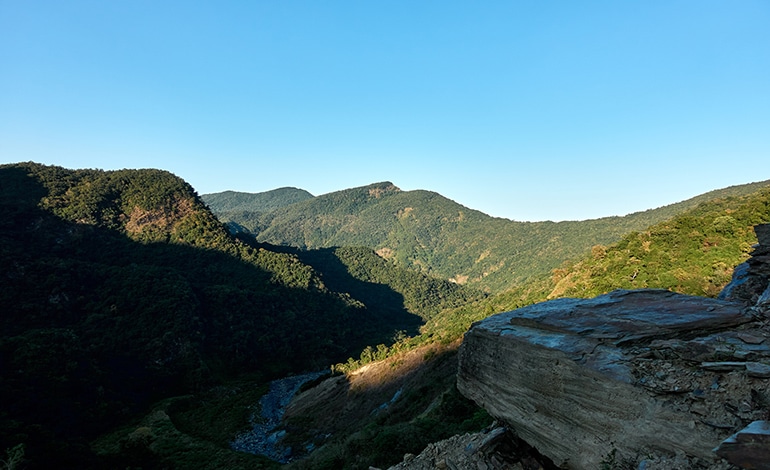 landscape picture of mountains with blue sky - river below - large boulder at bottom right