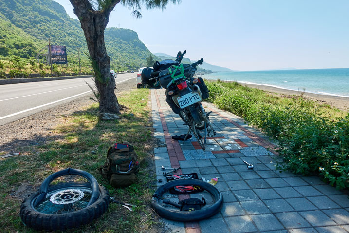 motorcycle on sidewalk without rear tire - ocean to the right - road to the left - tools and tire on the ground behind motorcycle