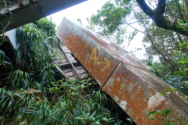 Rusted metal box-like structure that has fallen to the ground - an "A" and an arrow spray painted in yellow on it - some plants growing around it