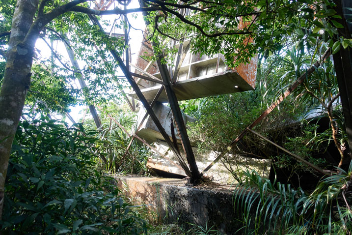 metal structure on top of concrete base - a few metal box-like structures have fallen and one suspended - some tree branches off to side