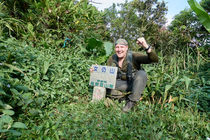 Me behind the triangulation stone in triumphant pose - holding sign with mountain name and elevation - trees behind - short grass in front