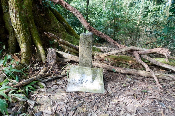 Stone marker with old sign in front of it - tree branches stacked behind it - WeiLiaoShan 尾寮山 trail