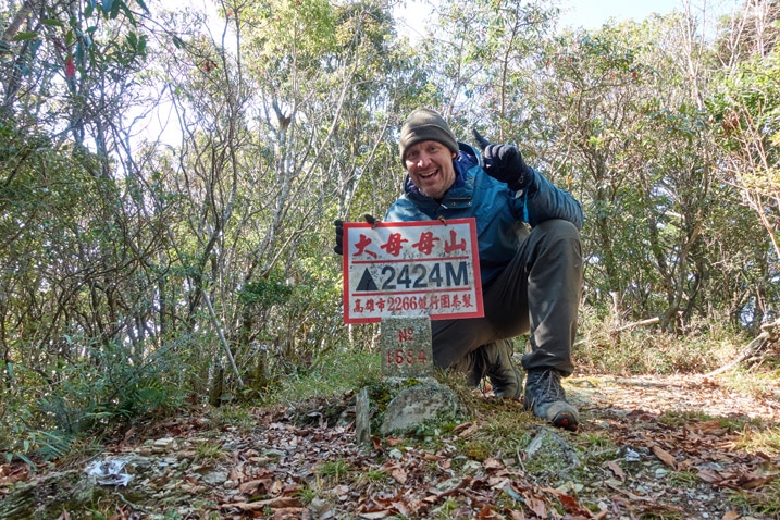 Man standing triumphantly behind 大母母山 Damumushan triangulation stone - holding sign - blue jacket