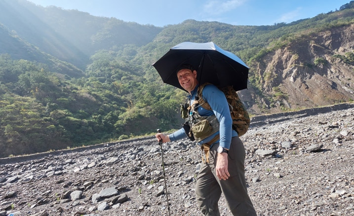 Man standing with umbrella over his head, holding a trekking pole - in rocky riverbed - mountains and blue sky