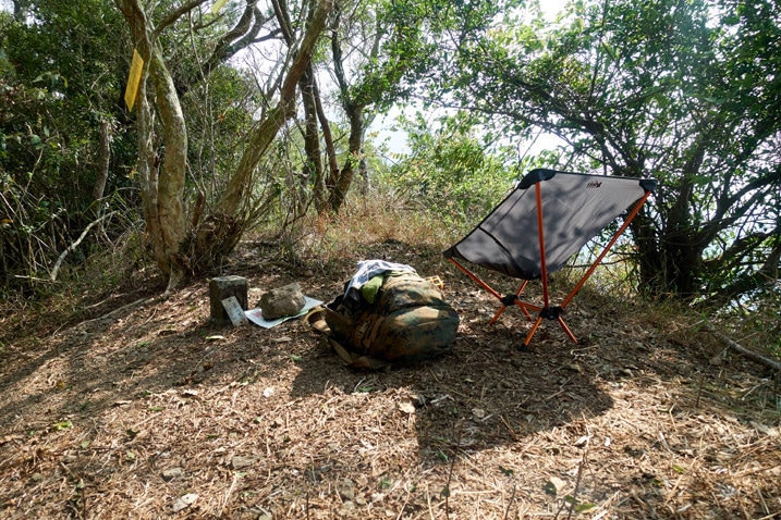 Camp chair, backpack and misc items on ground next to triangulation stone - trees all around
