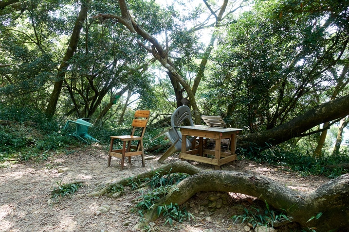 Wooden desk and two wooden chairs next to trail - trees behind them