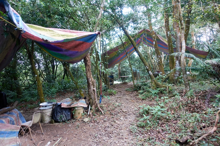 Two areas covered with colorful tarps - trees all around - tables, chairs and other items under tarps