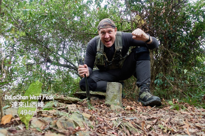 Man with machete kneeling triumphantly behind triangulation stone of DaLaiShan West Peak - 達來山西峰