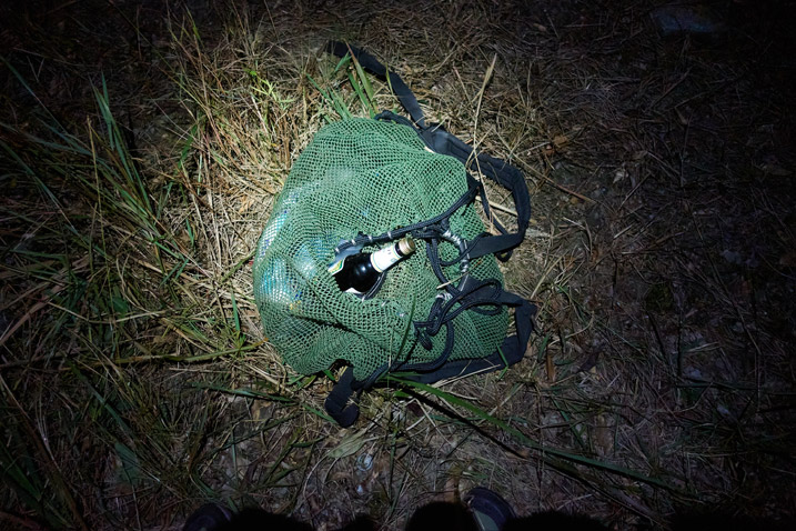 Green mesh bag on ground in the dark - flashlight illuminating - bottle sticking out of bag