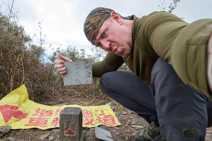 Man holding sign sideways while posing next to the BaCengBaMoShan 巴層巴墨山 triangulation stone 