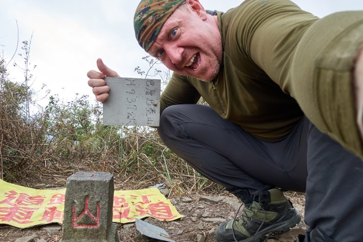 Man holding sign sideways while posing next to the BaCengBaMoShan 巴層巴墨山 triangulation stone 