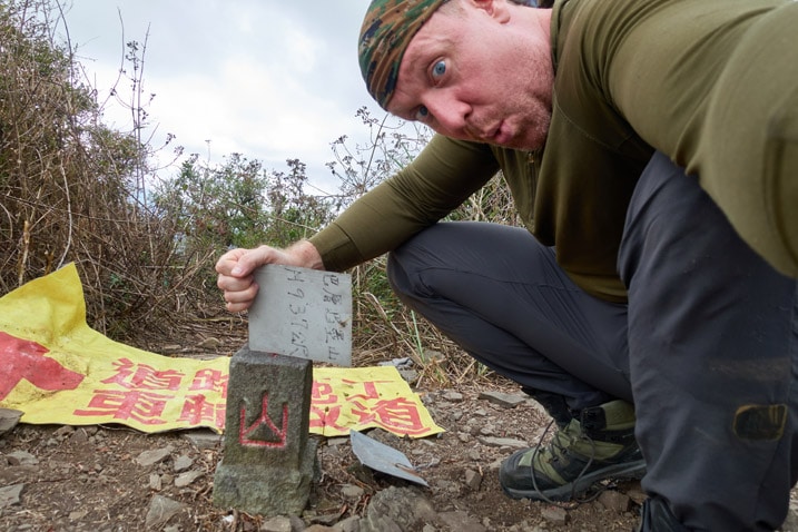 Man holding sign sideways while posing next to the BaCengBaMoShan 巴層巴墨山 triangulation stone 